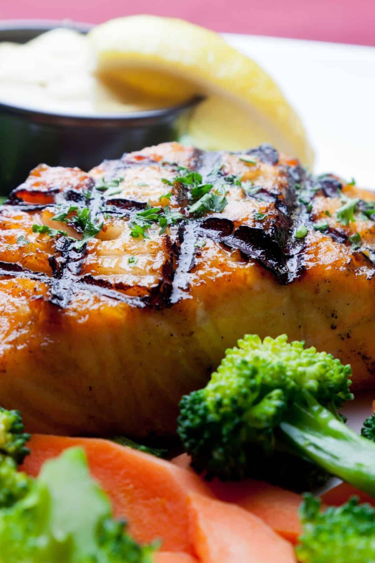 Grilled Salmon fillet with crosshatch grill marks, garnished with herbs, served with steamed broccoli, sliced carrots, lemon wedges, and a small cup of sauce in the background—a delightful Grilled Salmon dish that’s both vibrant and fresh.