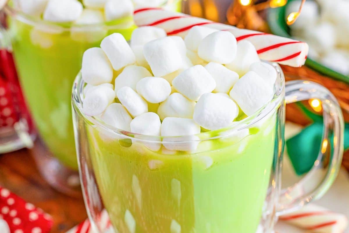 A clear mug filled with creamy hot chocolate in a vibrant green shade, topped with mini marshmallows and garnished with a striped peppermint stick—perfect inspiration for festive warm drinks.