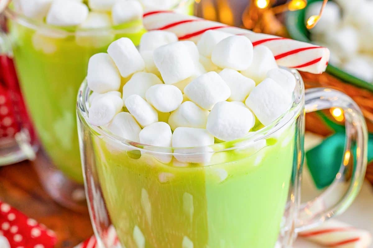 A mug of pastel green hot chocolate topped with marshmallows, with a candy cane on the side.