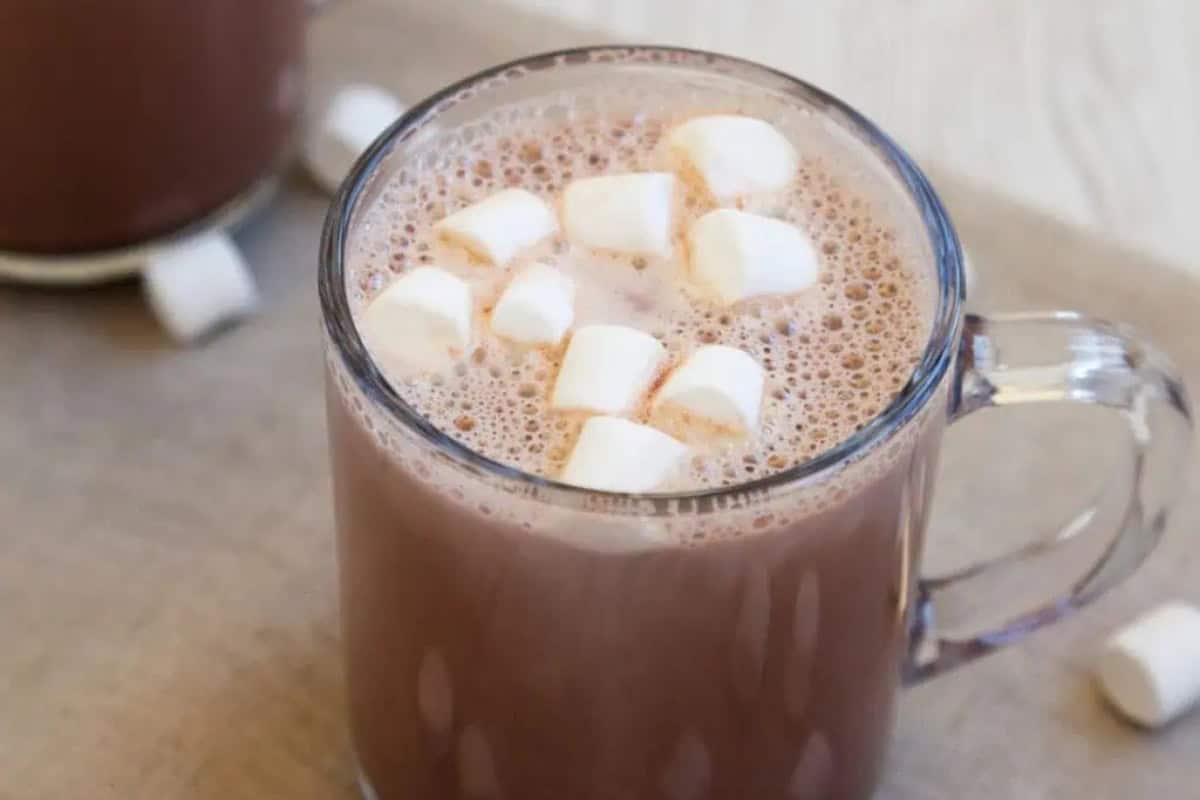 A clear glass mug filled with hot chocolate, topped with several small marshmallows, sits on a beige surface.