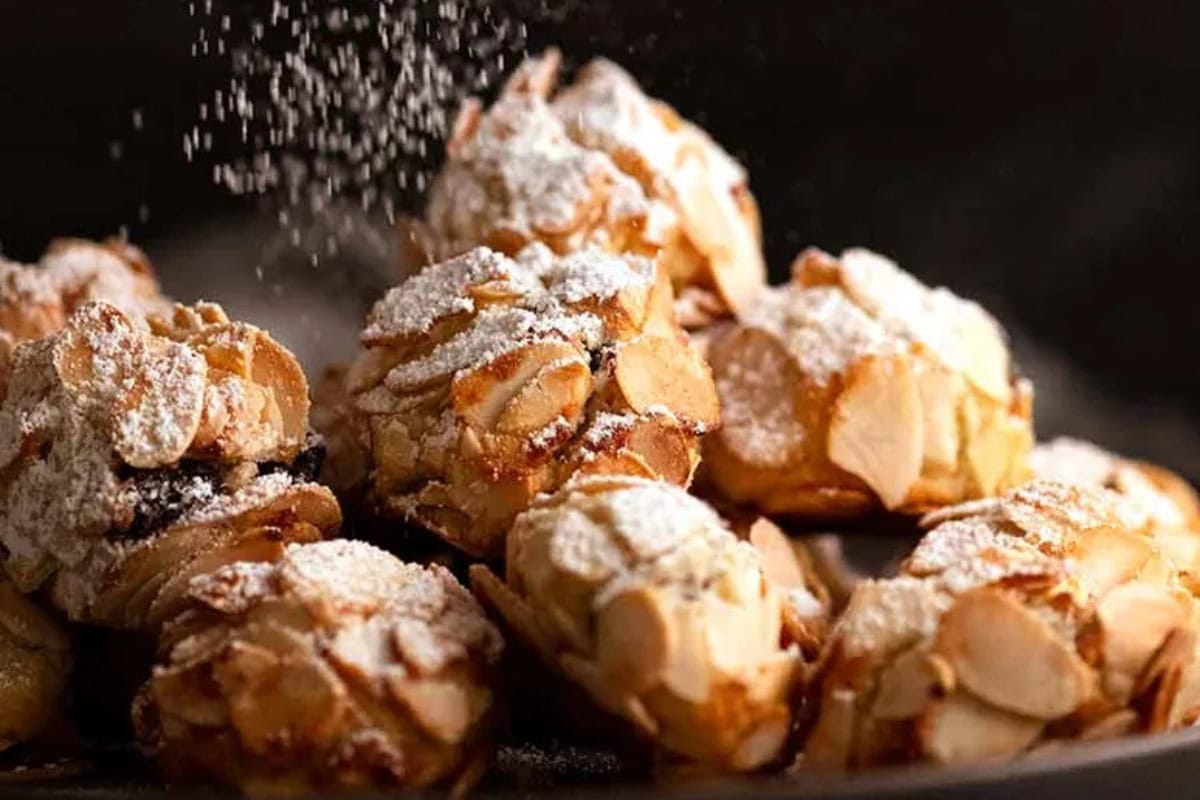 A close-up of several traditional Italian holiday cookies topped with sliced almonds and dusted with powdered sugar. Powdered sugar can be seen raining from above the cookies.