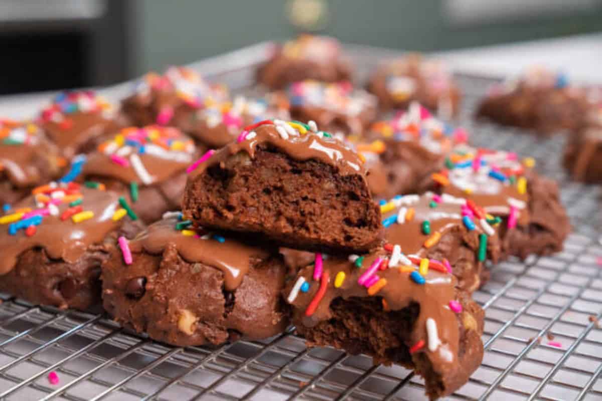 Chocolate meatball cookies topped with chocolate icing and colorful sprinkles, cooling on a wire rack, with one cookie broken in half to reveal the inside.