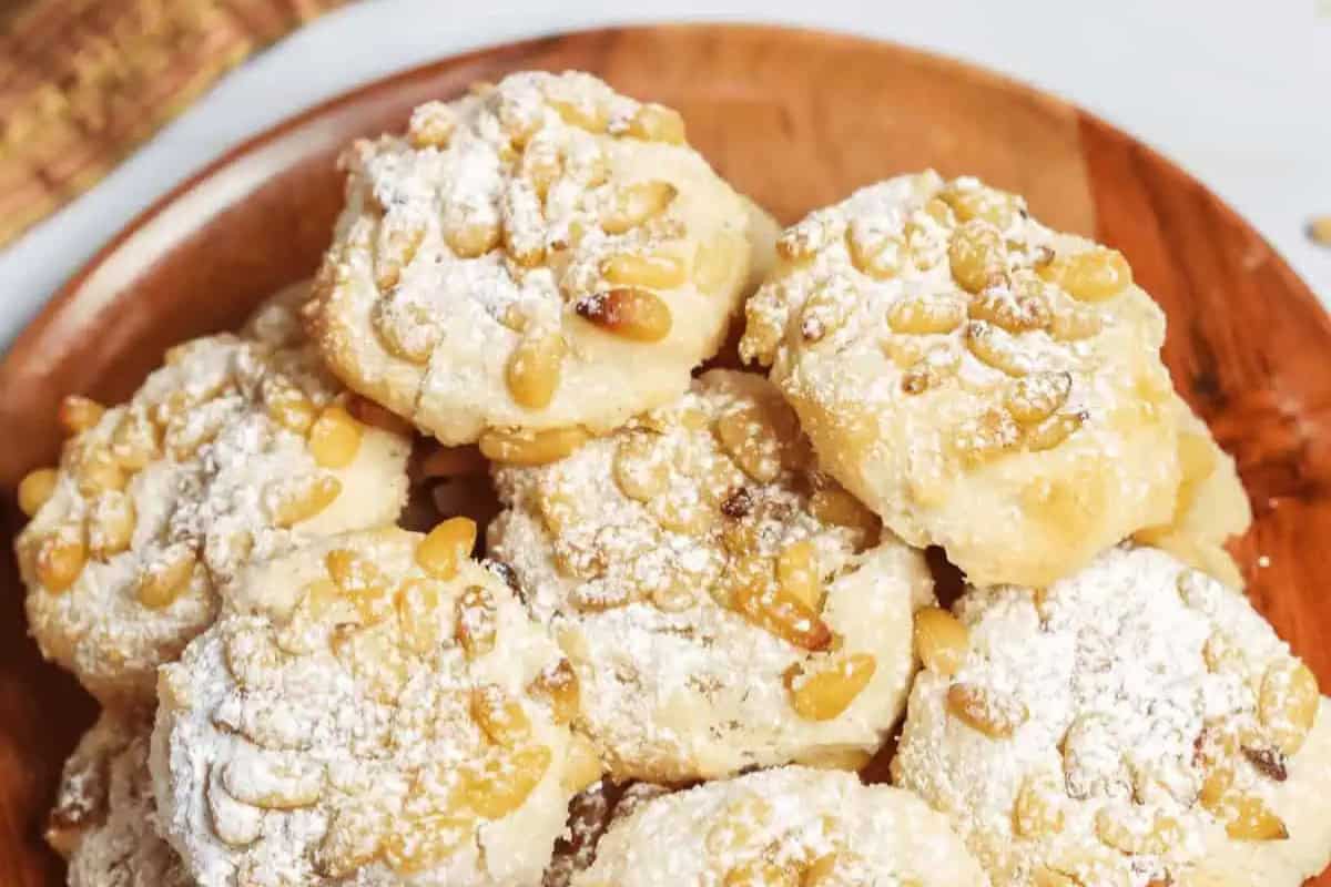 A wooden plate piled with traditional Italian cookies, topped with pine nuts and dusted with powdered sugar.