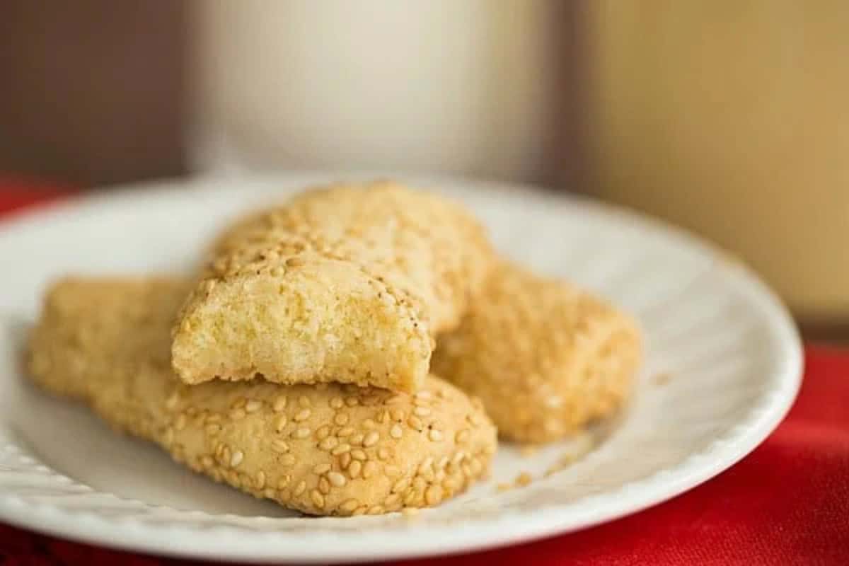 Sesame-coated traditional Italian cookies are stacked on a white plate, with one cookie broken in half to reveal the inside.