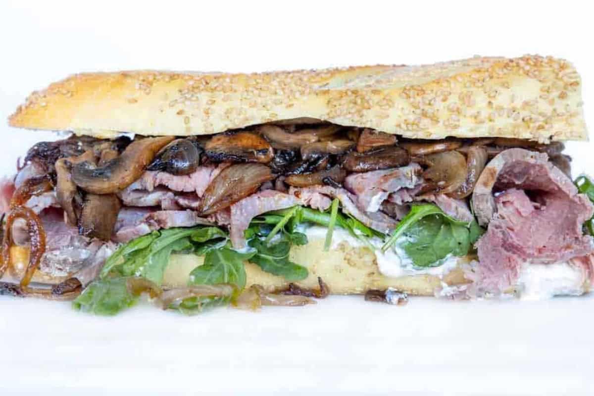 A sandwich on a sesame seed baguette filled with roast prime rib beef, sautéed mushrooms, arugula, caramelized onions, and a layer of white horseradish spread.