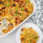 A serving of Creamy Chicken Noodle Casserole topped with crushed crackers, cheese, bacon, and chopped green onions on a plate next to a casserole dish with a checkered towel.