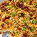A baked dish topped with crushed crackers, chopped bacon, and sliced green onions, with a serving scooped out from one corner.