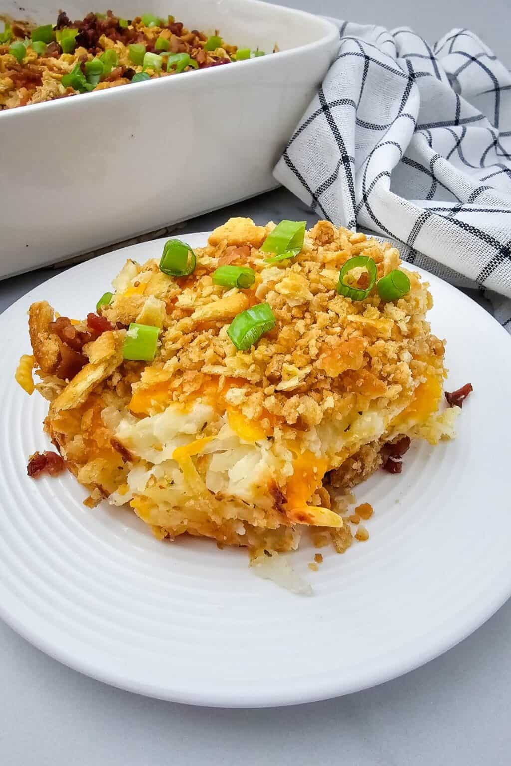 A serving of hash brown casserole, it is topped with crushed crackers and chopped green onions on a white plate, with a casserole dish and checkered towel in the background.