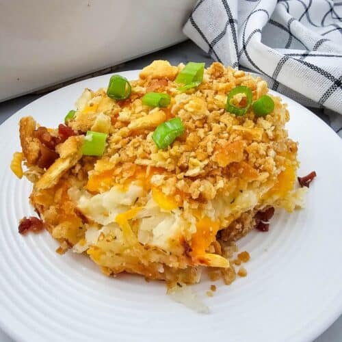 A serving of hash brown casserole, it is topped with crushed crackers and chopped green onions on a white plate, with a casserole dish and checkered towel in the background.