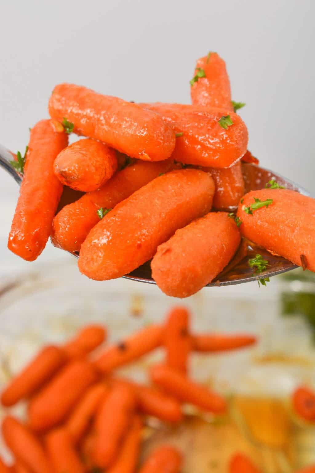A close-up of maple glazed carrots garnished with chopped herbs, held on a metal spoon above a bowl of more carrots.