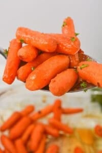A close-up of maple glazed carrots garnished with chopped herbs, held on a metal spoon above a bowl of more carrots.