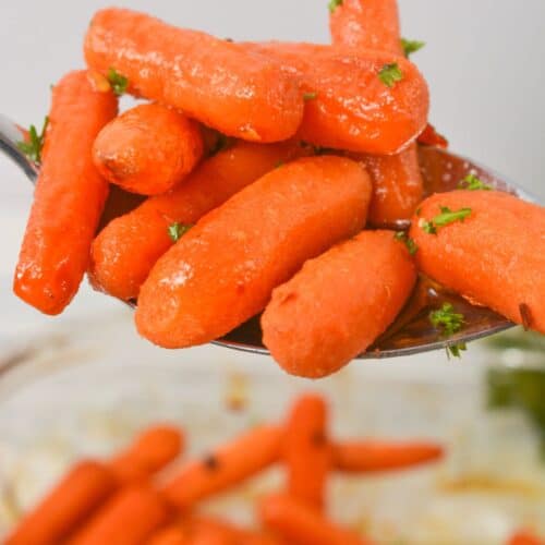 A close-up of maple glazed carrots garnished with chopped herbs, held on a metal spoon above a bowl of more carrots.
