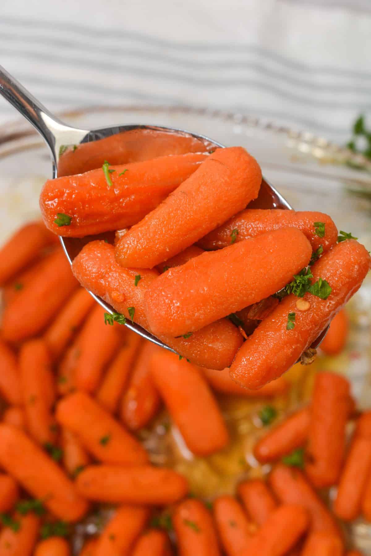 A spoon holds several cooked baby carrots garnished with chopped parsley, above a bowl filled with more maple glazed carrots and broth.