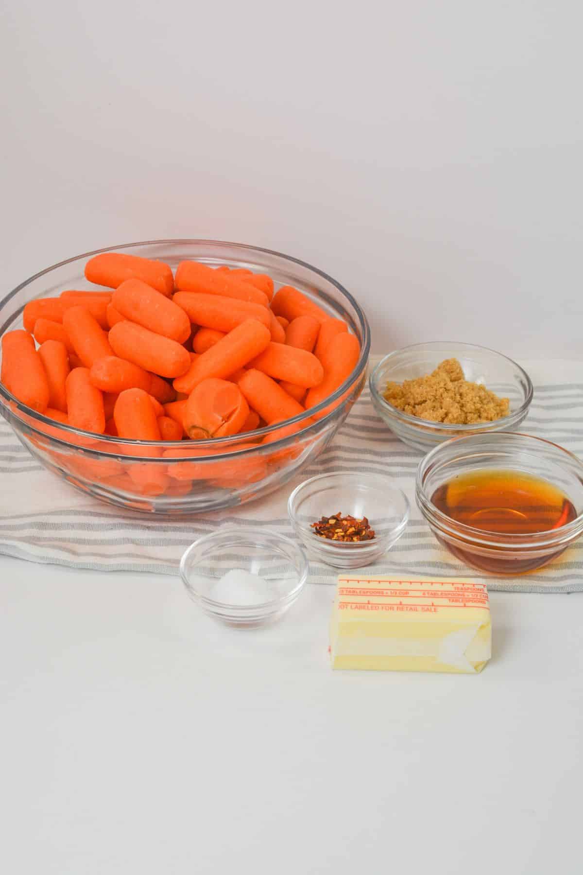 A glass bowl of baby carrots with small bowls of brown sugar, red pepper flakes, salt, honey, and a stick of butter on a striped cloth—everything you need to create delicious Maple Glazed Carrots.