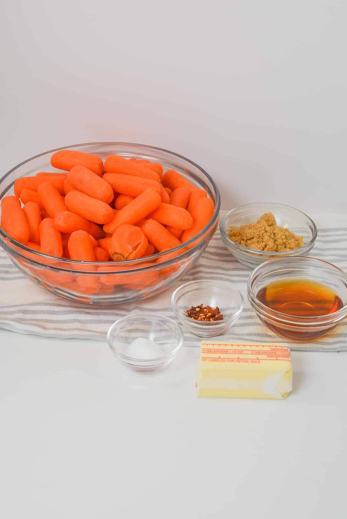 A bowl of baby carrots rests on a striped cloth, accompanied by small bowls of brown sugar, maple syrup, salt, crushed red pepper flakes, and a stick of butter—everything needed for delicious recipe.