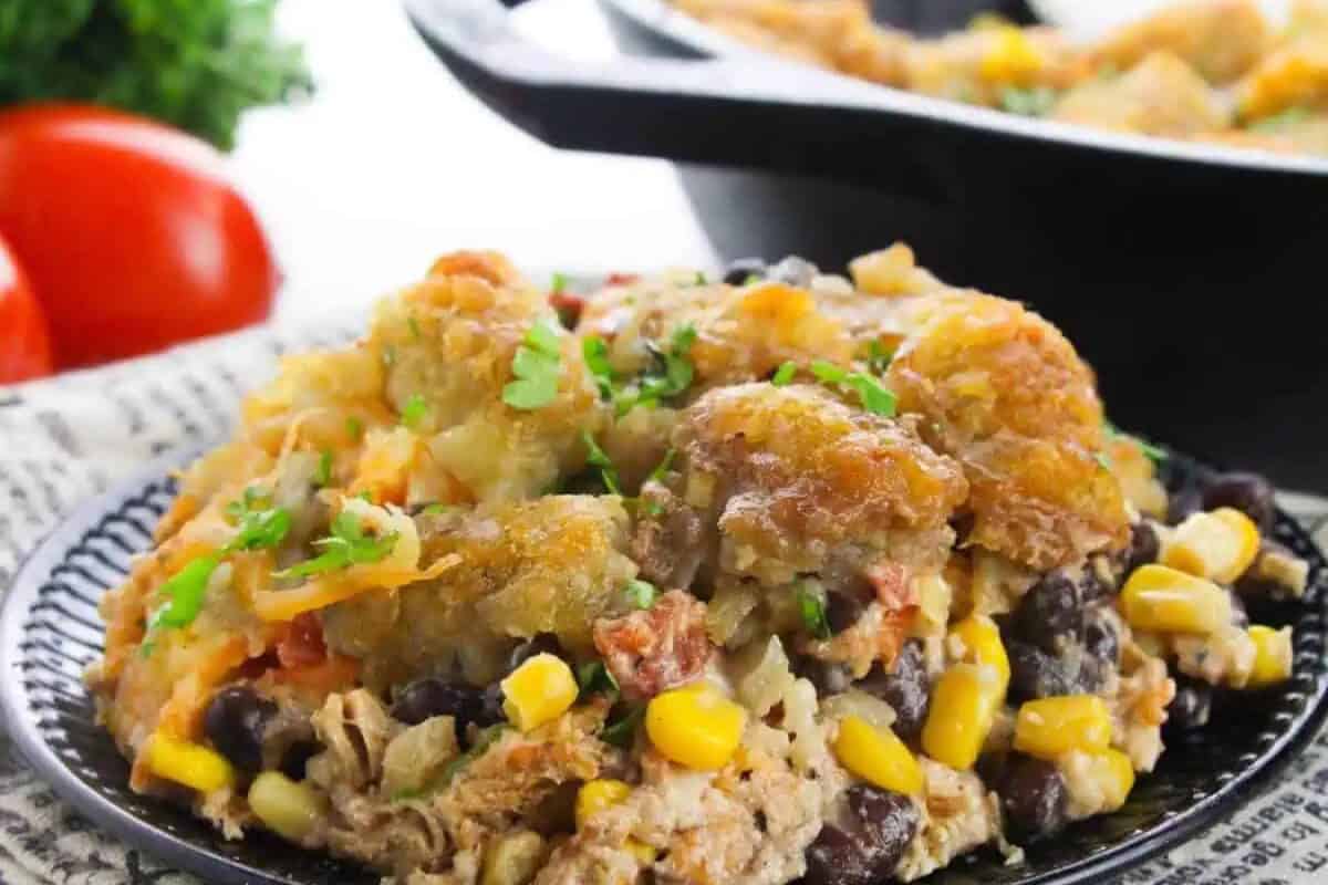 A serving of comfort food casserole featuring tater tots, black beans, corn, shredded meat, and herbs on a black plate, with a pan of chicken casseroles and vegetables in the background.