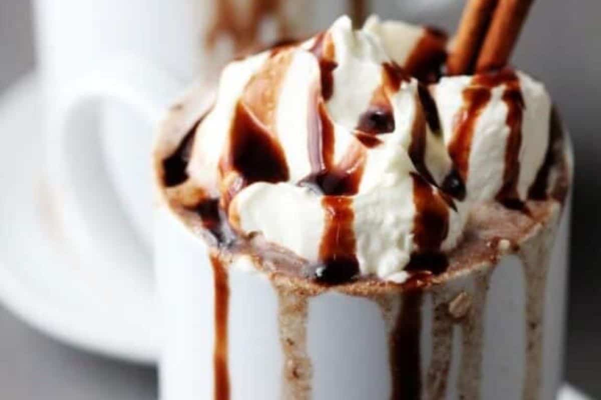 A white mug filled with rich hot chocolate, topped with whipped cream, chocolate syrup, and two cinnamon sticks, with another mug blurred in the background.