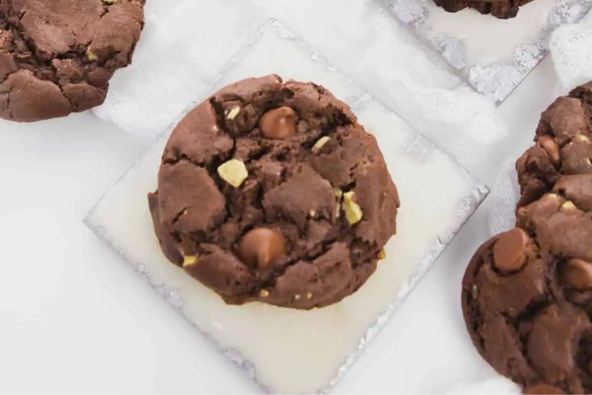 A chocolate cookie with chocolate chips and Andes Candies pieces sits on a white square tile, perfect for Cozy Christmas gatherings, with other cookies visible nearby.