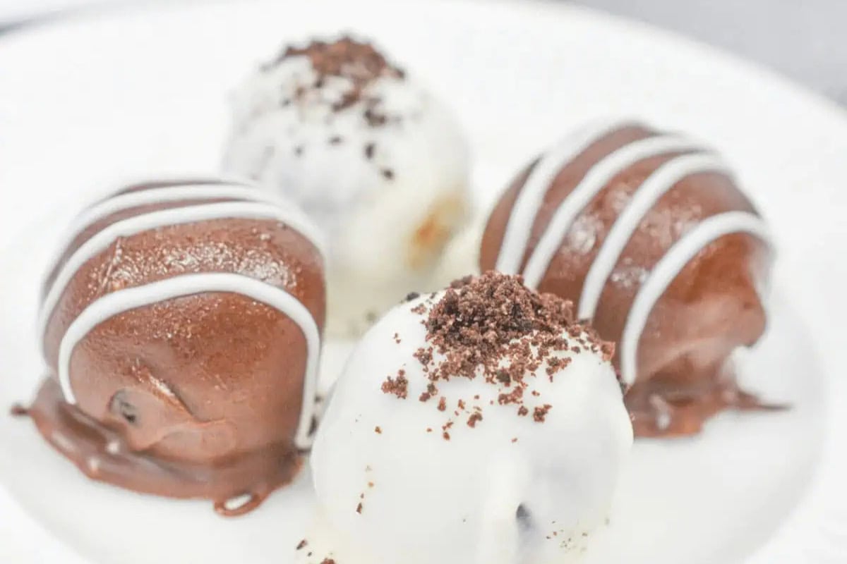 Four round chocolate Oreo truffles on a white plate, two coated in dark chocolate and two in white chocolate, each decorated with white icing or chocolate crumbs.