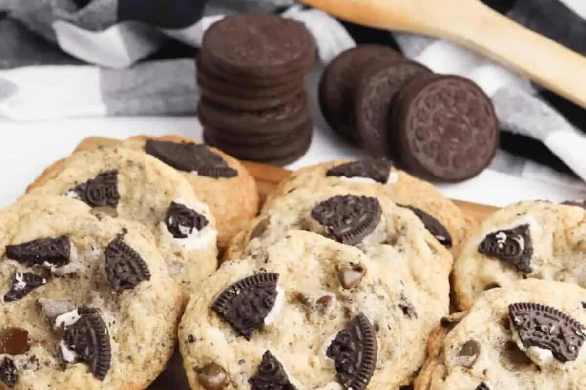 Chocolate chip cookies with pieces of chocolate sandwich Oreo cookies on top are arranged on a wooden surface. Whole sandwich cookies sit in the background.