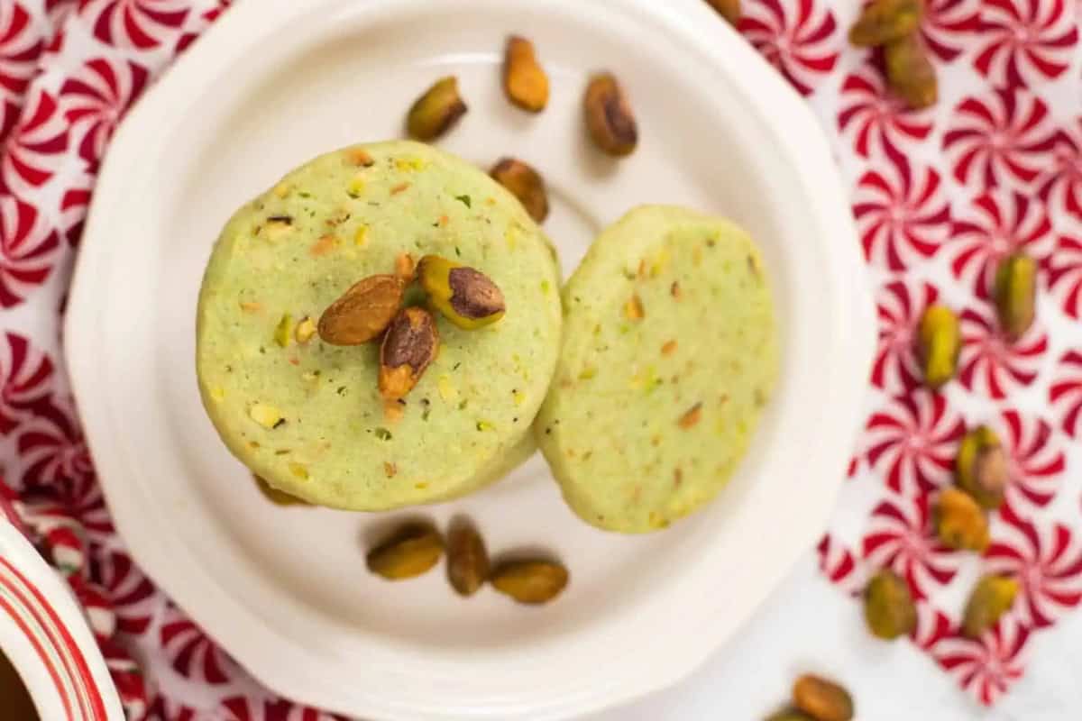 A birds-eye view of a stack of round traditional Italian pistachio cookies with an additional cookie leaning sideways off of the stack sit on a white plate, garnished with whole pistachios, with more pistachios scattered on a red and white peppermint patterned surface.