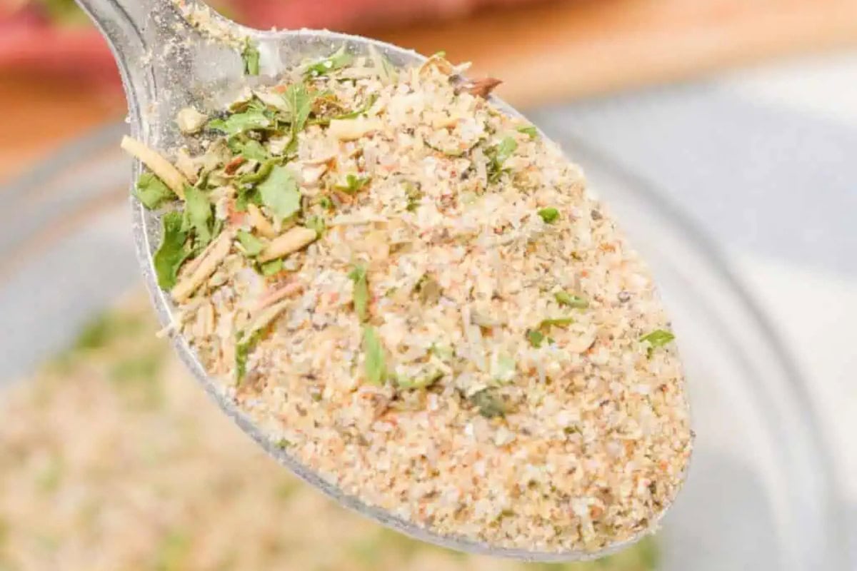 A close-up of a spoon holding a blend of dried herbs and spices, with green, brown, and beige particles visible.