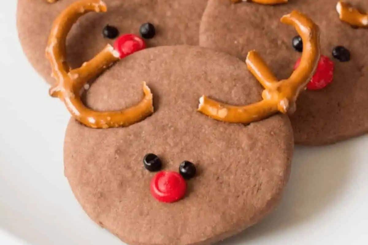 Cute reindeer-shaped cookies with pretzel antlers and candy eyes and nose, sitting on a white tray.