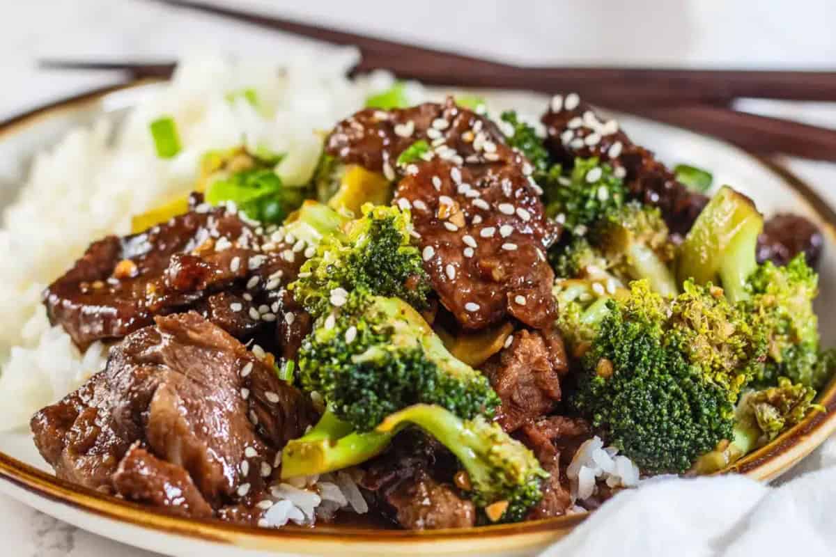 A plate of prime rib beef and broccoli stir-fry served over white rice, topped with sesame seeds, with chopsticks in the background