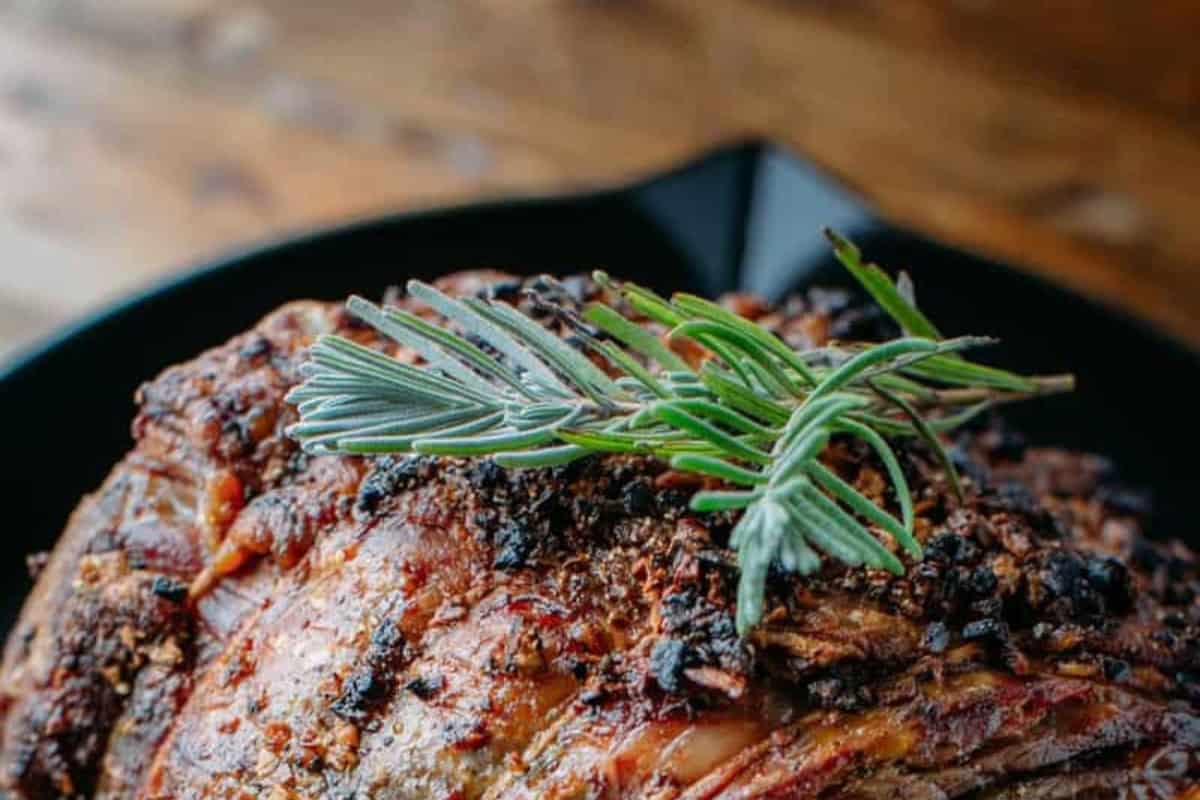 A close-up of a roasted prime rib meat dish,topped with a sprig of fresh rosemary and served in a dark pan on a wooden surface.