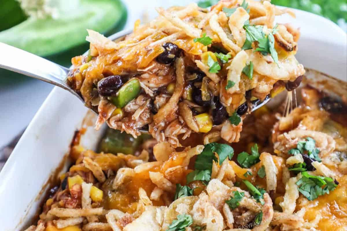 A spoonful of comfort food casserole with black beans, corn, shredded cheese, and crispy fried onions, garnished with chopped cilantro, held above a baking dish—perfect for fans of hearty chicken casseroles.