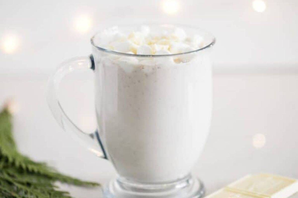 A clear glass mug filled with a creamy hot cocoa topped with mini marshmallows, placed on a white surface with blurred lights in the background.