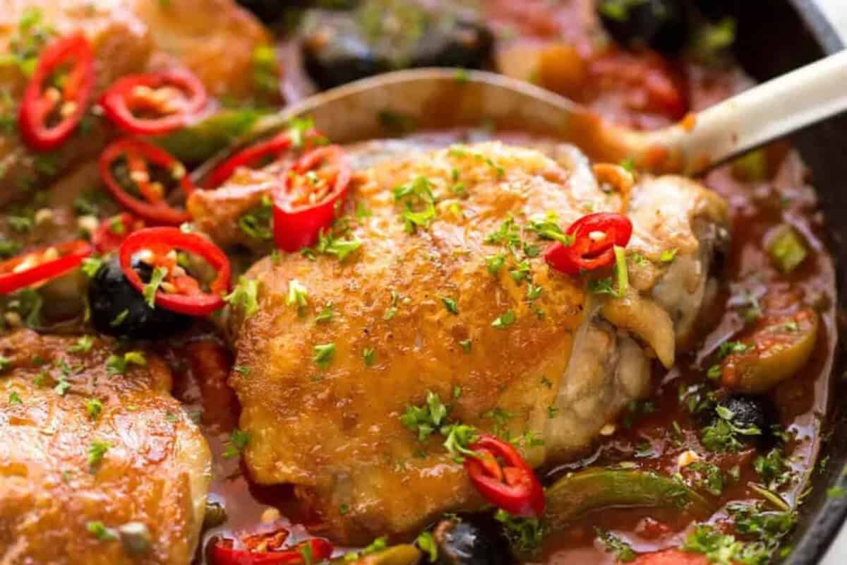 A cooked chicken thigh in tomato-based sauce, reminiscent of classic comfort food and hearty chicken casseroles, garnished with sliced red chili and chopped herbs, served with vegetables and olives.
