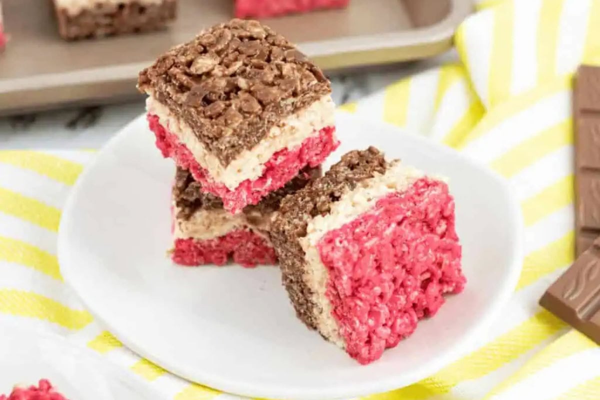 Three layered Rice Krispies treats with chocolate, white, and pink layers are stacked on a white plate. A baking tray and chocolate bar sit in the background.