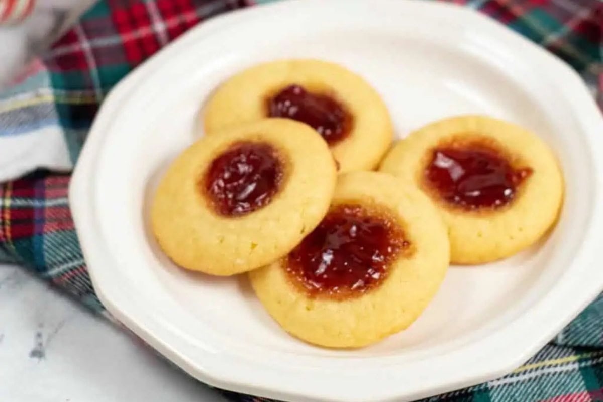 Four round thumbprint cookies filled with red strawberry jam are arranged on a white plate, set atop a plaid cloth background.