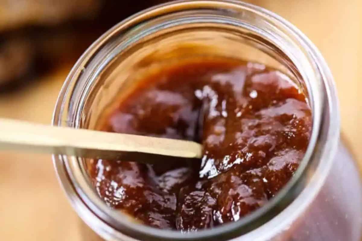 A close-up view of a glass jar filled with sweet and spicy BBQ sauce, with a wooden stick partially submerged in the contents.