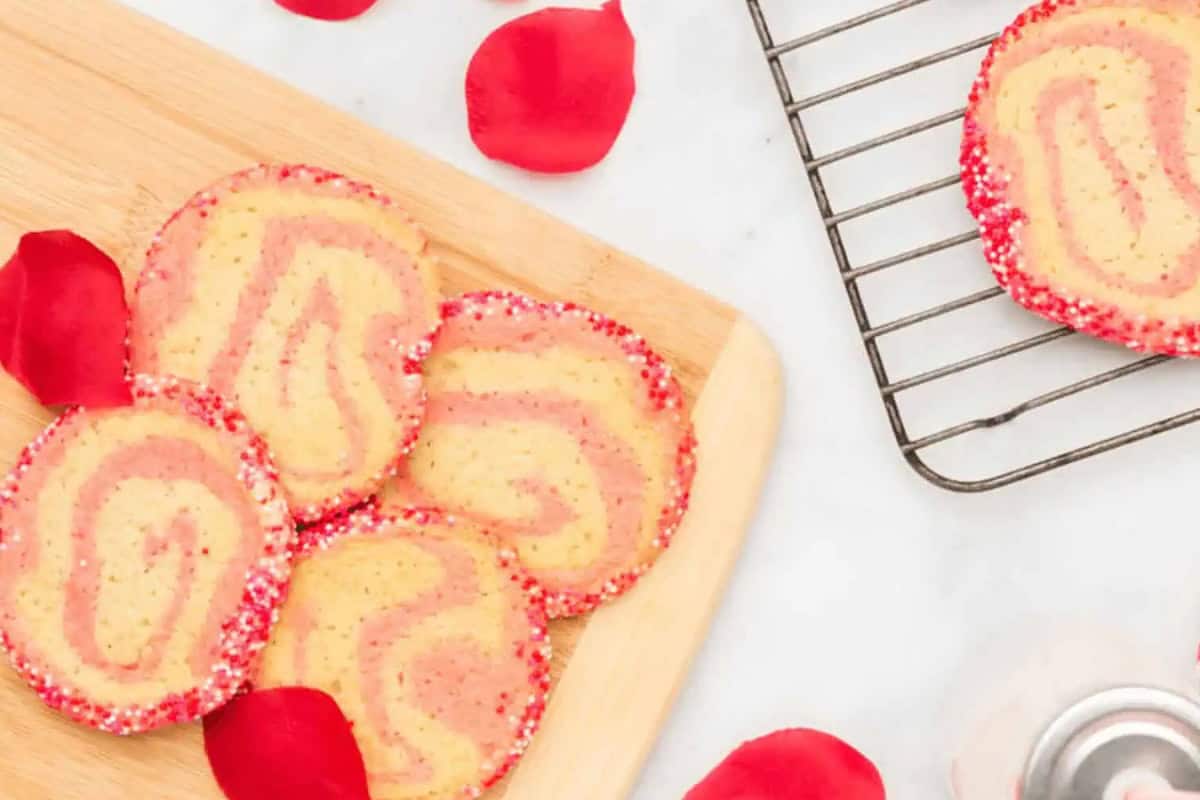 Five pink and yellow swirl Christmas cookies with red sprinkles sit on a wooden board next to a cooling rack and scattered red rose petals.