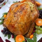 A Turkey in an Oven Bag stuffed with bread stuffing is served on a platter with grapes, cranberries, orange slices, carrots, and herbs on a decorated table.