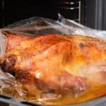A whole roasted duck with crispy skin is cooking in an oven, wrapped in a clear roasting bag on a baking tray, similar to preparing turkey in an oven bag for juicy, flavorful results.
