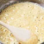 A wooden spoon stirs a bubbly, thick yellow batter in a metal saucepan, much like making Turkey Gravy from the Drippings after a holiday meal.