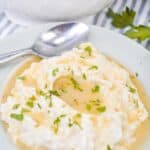 A plate of mashed potatoes topped with brown gravy made from Turkey Gravy without Turkey Drippings, garnished with chopped parsley, with a spoon resting on the plate and a bowl of gravy in the background.