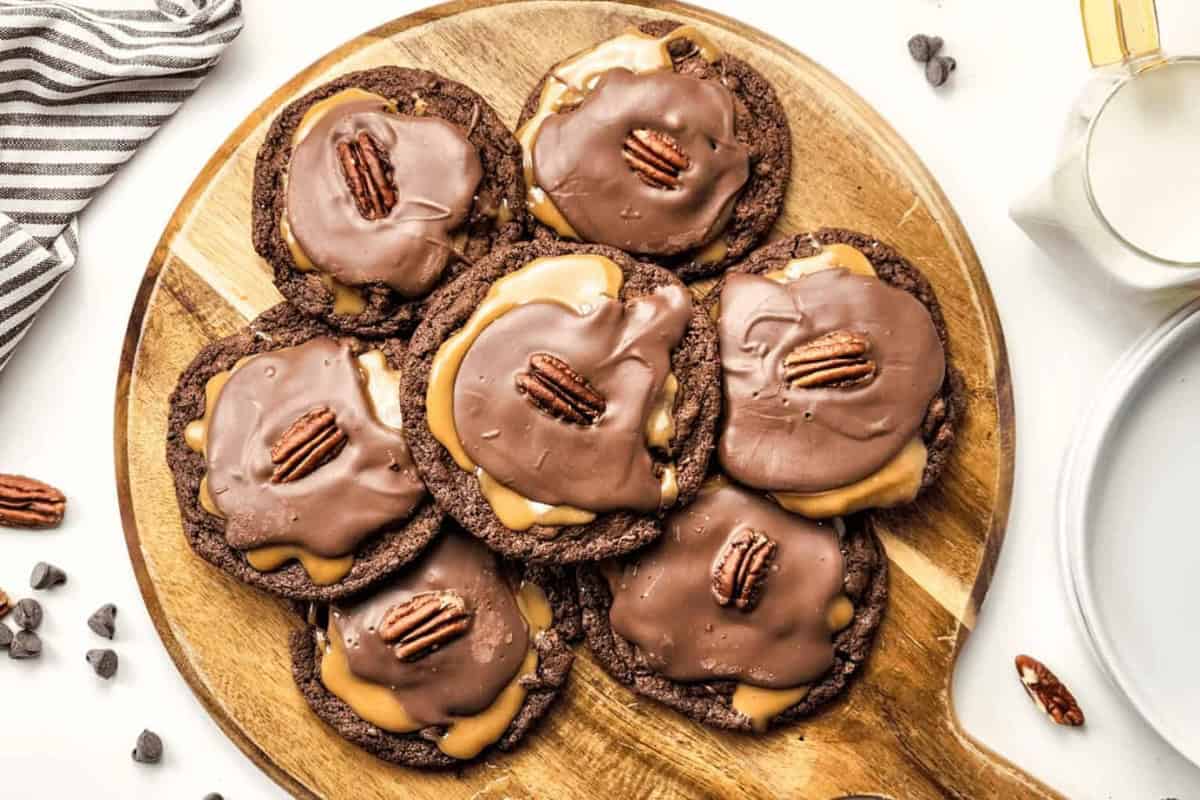 A group of chocolate cookies covered with chocolate frosting and caramel topped with pecans on a wooden board.