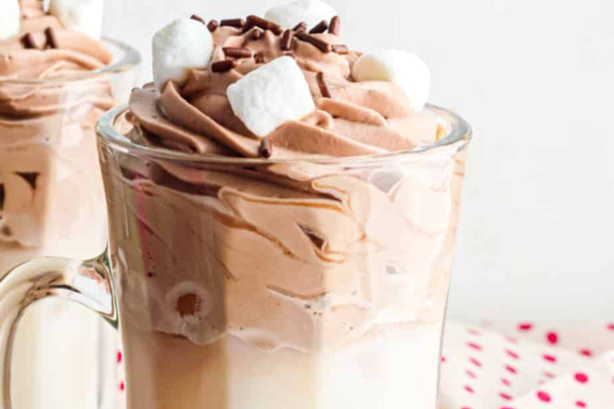 A glass mug of creamy hot whipped chocolate topped with mini marshmallows and chocolate sprinkles.