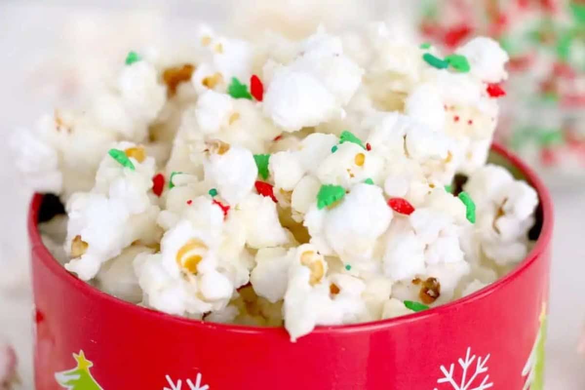 A red tin decorated with white snowflakes and Christmas trees is filled with white chocolate popcorn topped with red and green sprinkles.