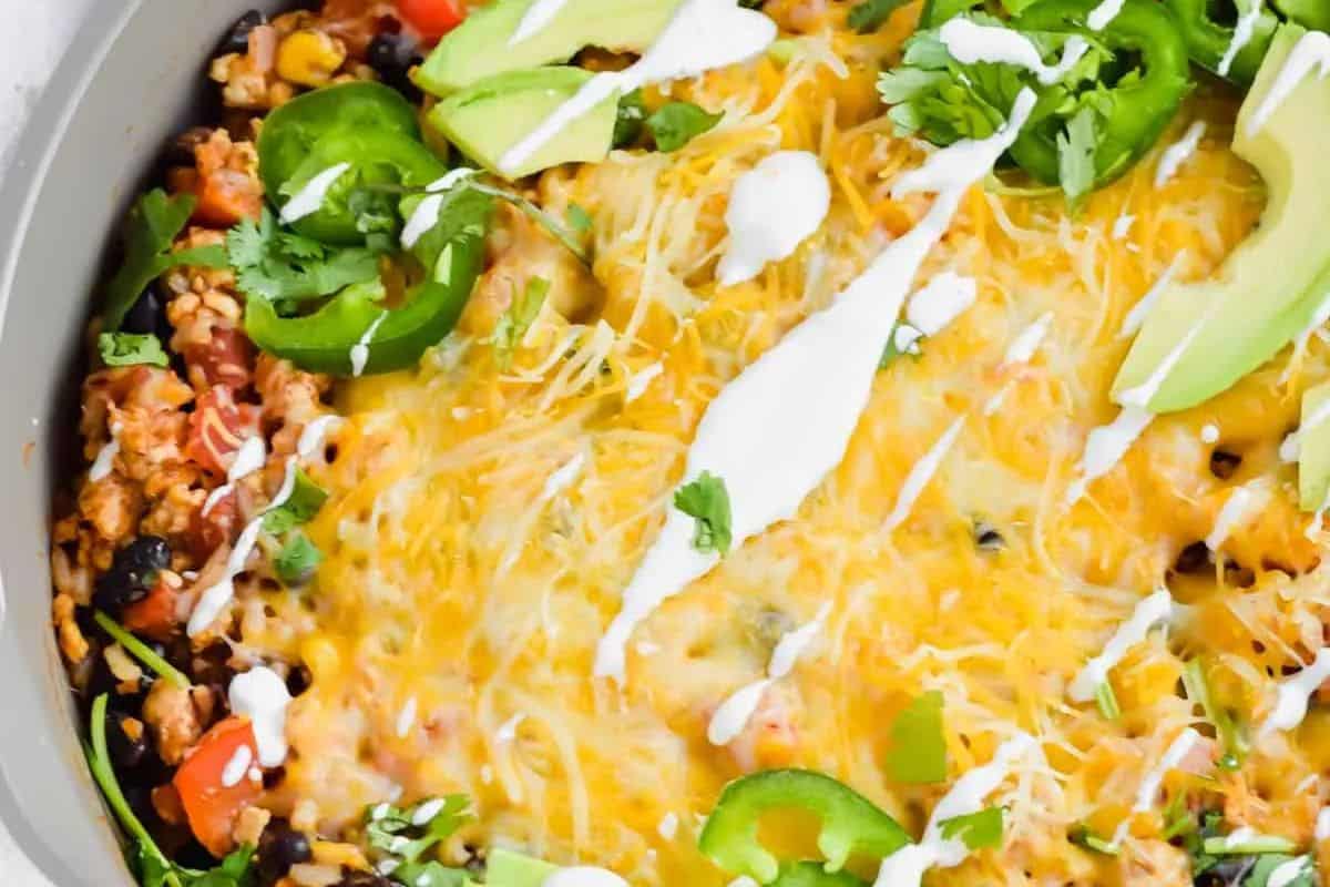 Close-up of a cheesy Mexican-inspired casserole—perfect for cooler weather—topped with sliced avocado, jalapeños, cilantro, and drizzled sour cream. A must-try for fans of good casserole recipes.