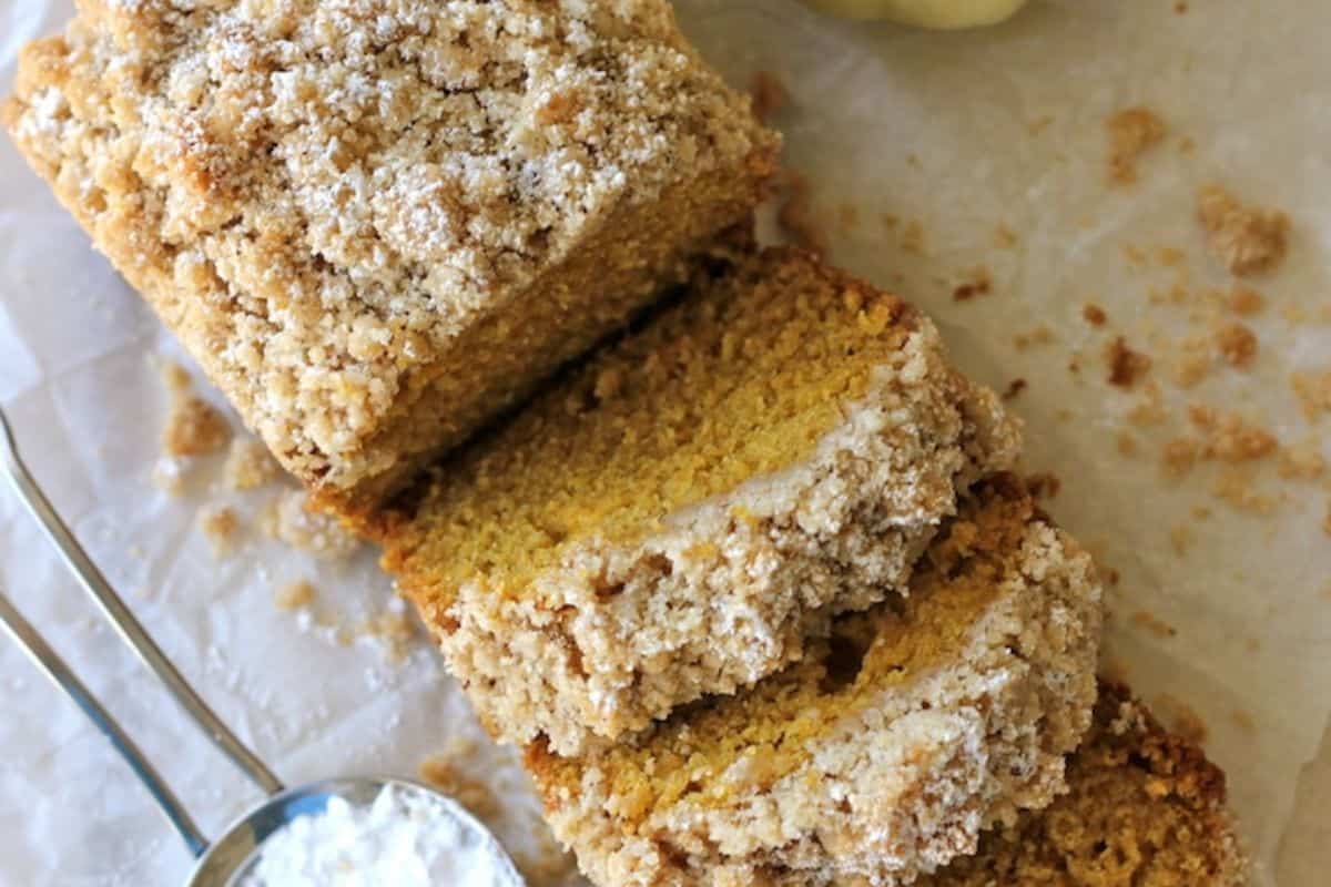 A loaf of crumb-topped bread is partially sliced on parchment paper, evoking fall vibes, with a metal measuring spoon containing powdered sugar nearby.