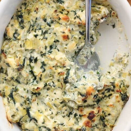 A white oval dish filled with baked spinach and artichoke dip, with a serving spoon and a portion removed, showing the creamy texture—a perfect choice for easy appetizers at your next gathering.