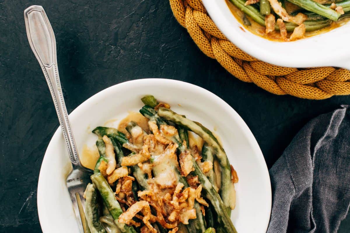 A bowl of green bean casserole topped with crispy fried onions, with a fork on the side. Perfect for cooler weather, this good casserole sits beside a larger serving dish and a woven mat that are partially visible nearby.