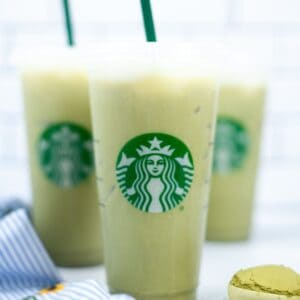Three iced matcha lattes in clear plastic cups with green straws, prominently featuring the Starbucks logo, rest on a white surface. Nearby, a striped cloth and a delicate macaron complete the charming scene.