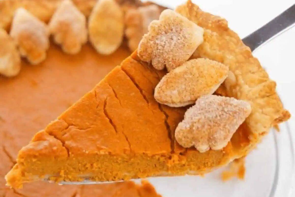 A slice of pumpkin pie with a flaky crust and leaf-shaped pastry decorations is being lifted from the whole pie, capturing the essence of cozy fall and classic pumpkin desserts.