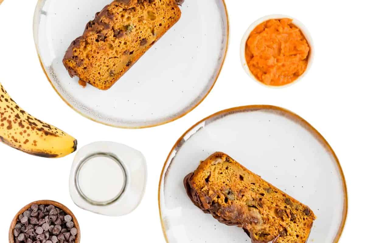 Two slices of pumpkin banana bread on plates, surrounded by a banana, chocolate chips, a small bowl of pumpkin puree, and a jug of milk on a white background—perfect for enjoying cozy fall vibes and pumpkin desserts.